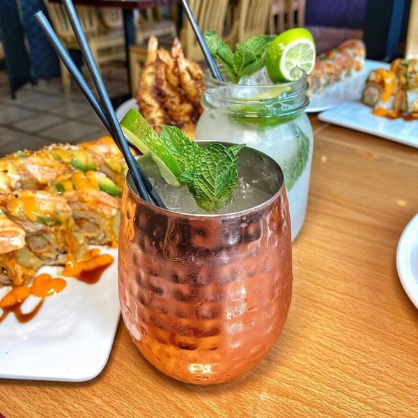 Two mojitos, one in a copper mug, with sushi rolls and fried shrimp on a wooden table.