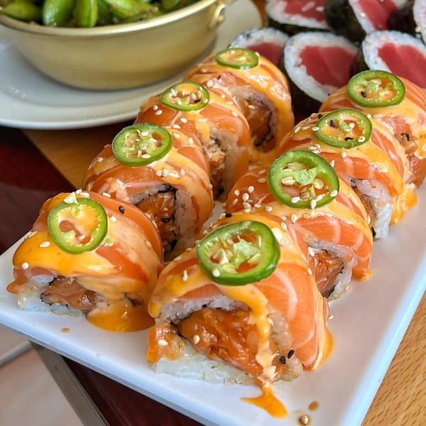 Tengoku Sushi's salmon sushi rolls with spicy sauce, sesame seeds, and jalapeño slices on a white plate.