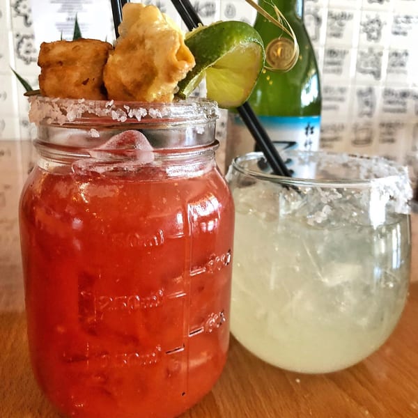 Two salt-rimmed cocktails, one red in a mason jar with fried garnishes, and one clear.