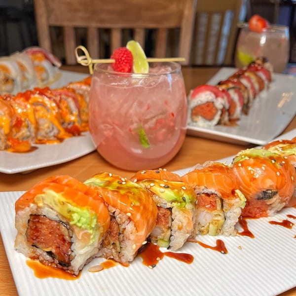 Plates of colorful sushi rolls and pink cocktails with fruit garnishes on a wooden table.