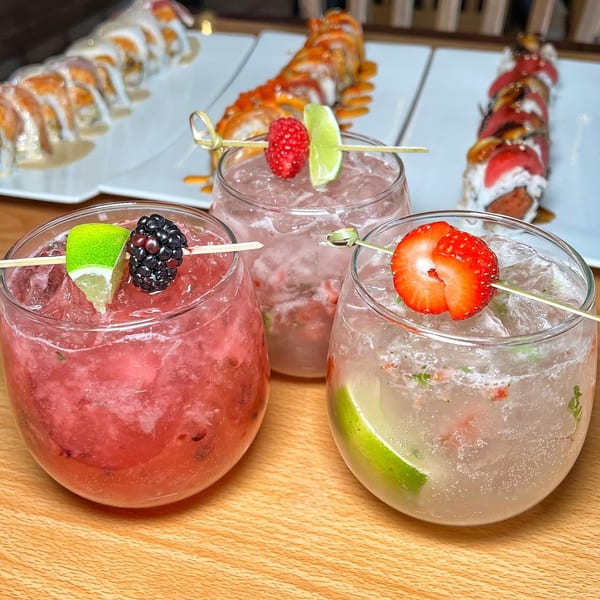 Three colorful berry cocktails with lime garnishes and sushi rolls in the background at Tengoku Sushi.