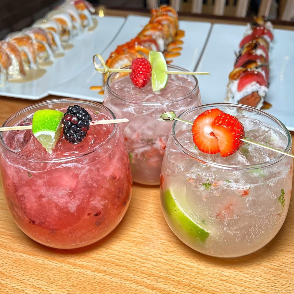 Three colorful berry cocktails with lime garnishes and sushi rolls in the background at Tengoku Sushi.